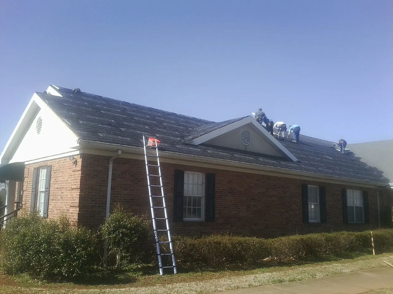 Crew tearing shingles off a brick ranch roof with a ladder set — Emergency Roof Repair work by our crew in Grass Lake