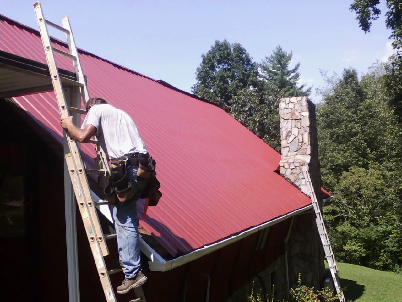 Roofer on a ladder beside a red metal roof and stone chimney — Soffit & Fascia Repair by Roofing Brothers in Grass Lake